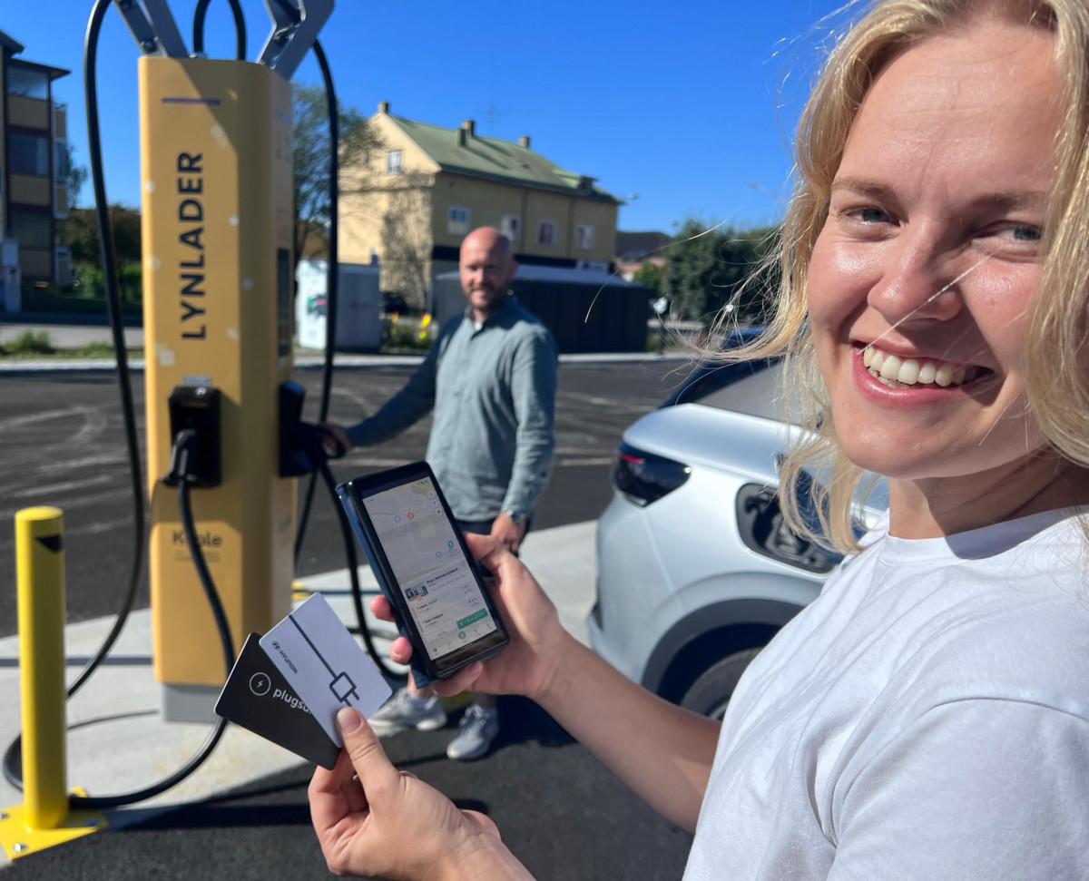 The image shows a cheerful woman in the foreground holding a payment card and a smartphone with an open app. She is looking towards the camera with a broad smile. In the background, slightly blurred, a man stands next to a white electric car charging at a yellow charging station marked 'LYNLADER'. The scene takes place in a parking lot with urban surroundings in the background.