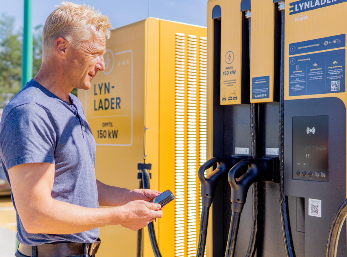 A man stands at an electric vehicle charging station, closely examining his smartphone. The charging station is branded 'LYNLADER' and indicates a 150 kW charging capability. The station is a bold yellow color, with two charging cables hanging ready for use. The man, dressed in casual clothing, seems engaged with the charging process or possibly an app related to the service. The focus on the man suggests a user-friendly and accessible electric vehicle infrastructure.